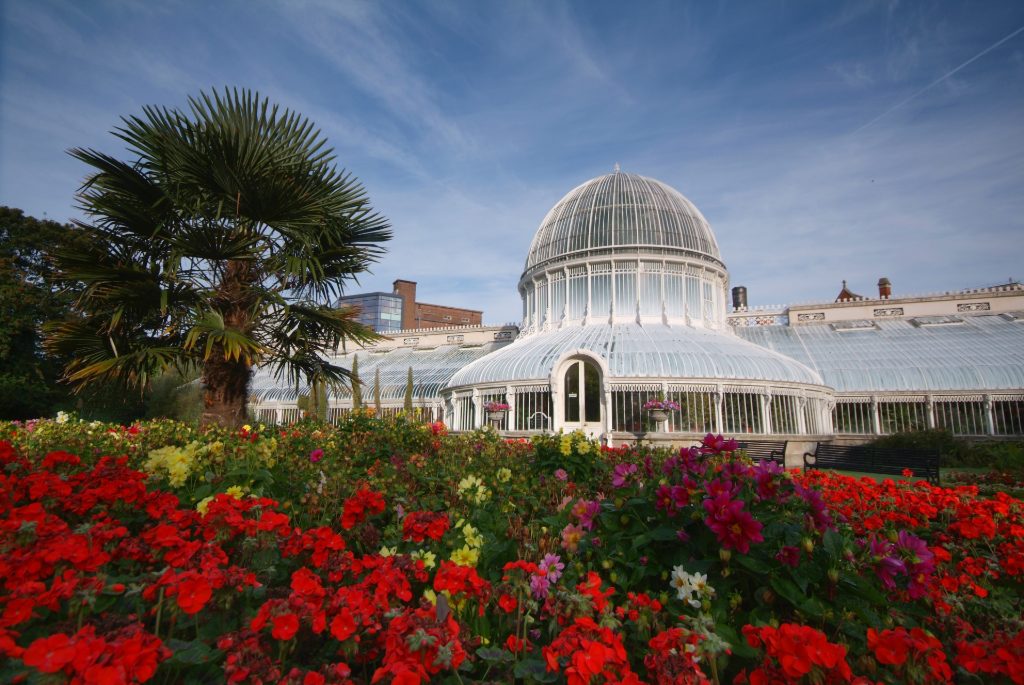 Victorian glasshouse framed by palm tree and vivid red blooms in summer sunshine.