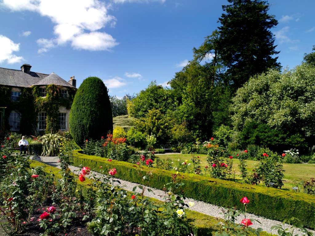 Formal rose garden with manicured hedges and historic house under blue skies.