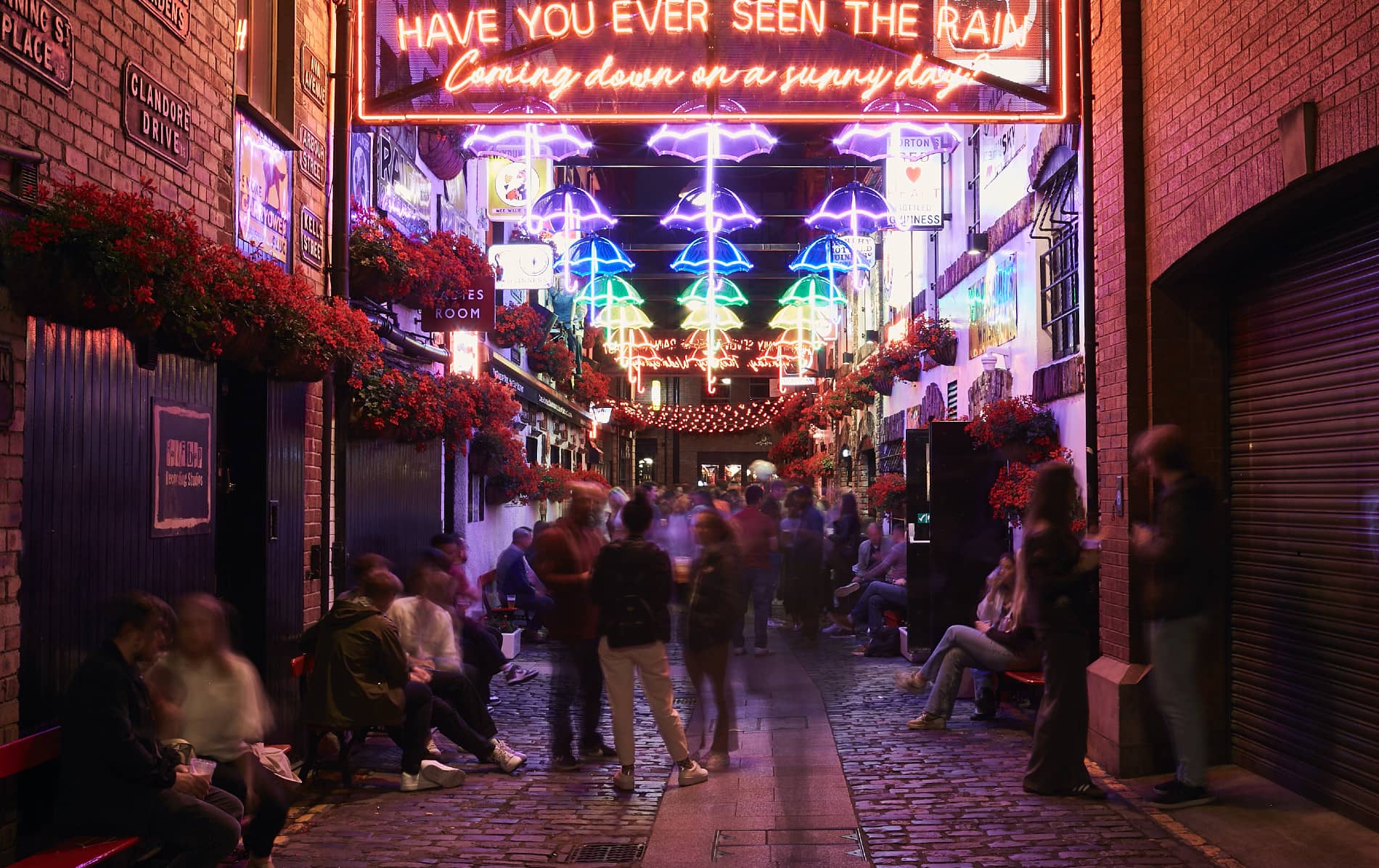 Cathedral Quarter alley in Belfast lit by neon umbrellas and signs, bustling with evening crowds.