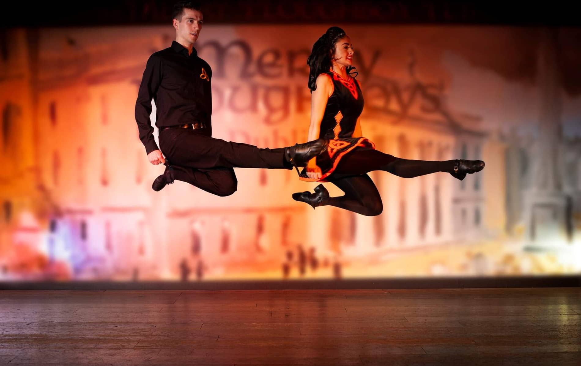 Irish dancers in black and red costumes leap mid-air during a traditional dance performance.
