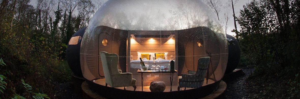 Luxury bubble dome accommodation in a forest clearing at Finn Lough, County Fermanagh, lit warmly at dusk.