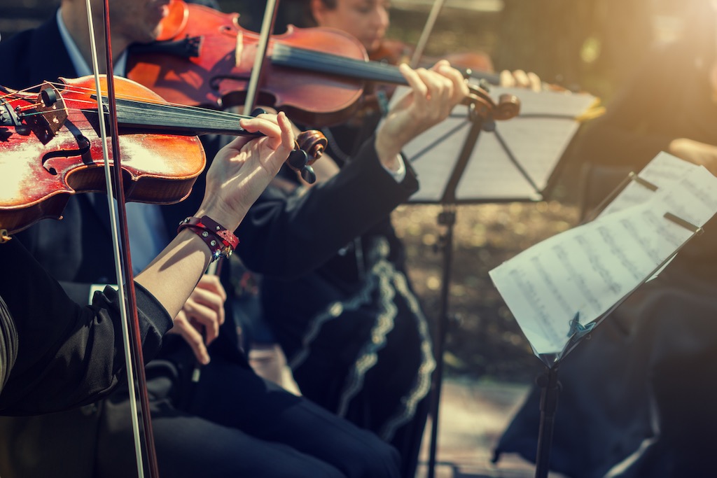 Close-up of musicians playing violins outdoors with sheet music stands.