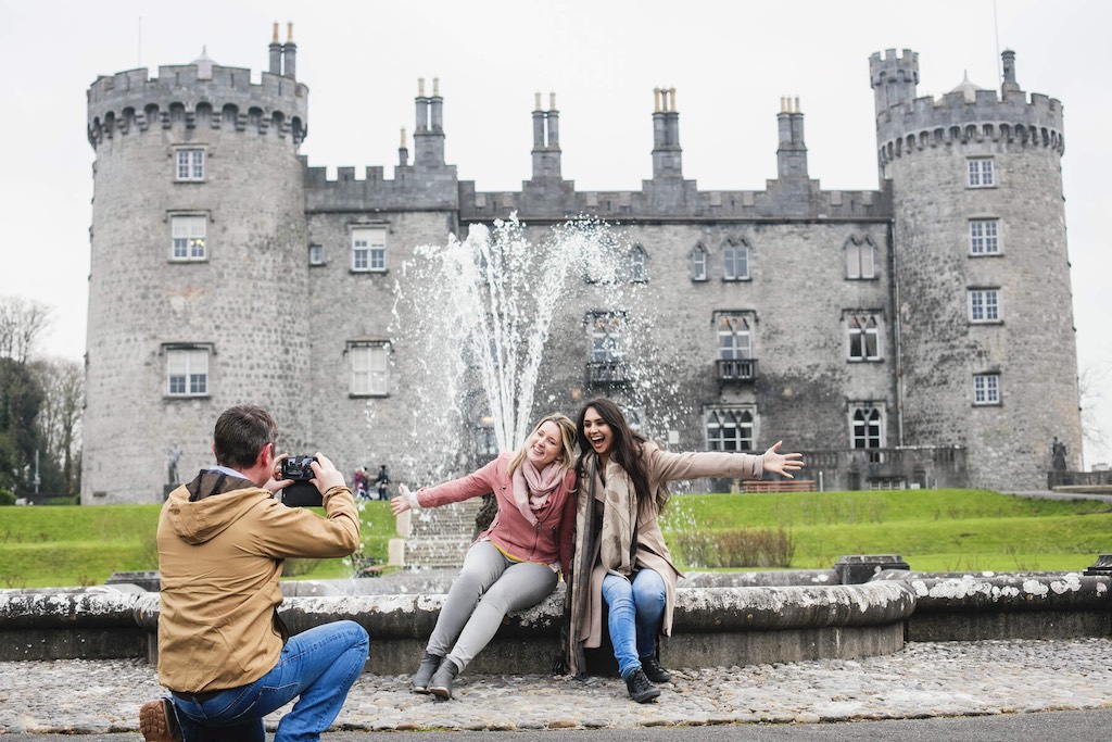 Tourists posing by the fountain in front of Kilkenny Castle on a cloudy day.
