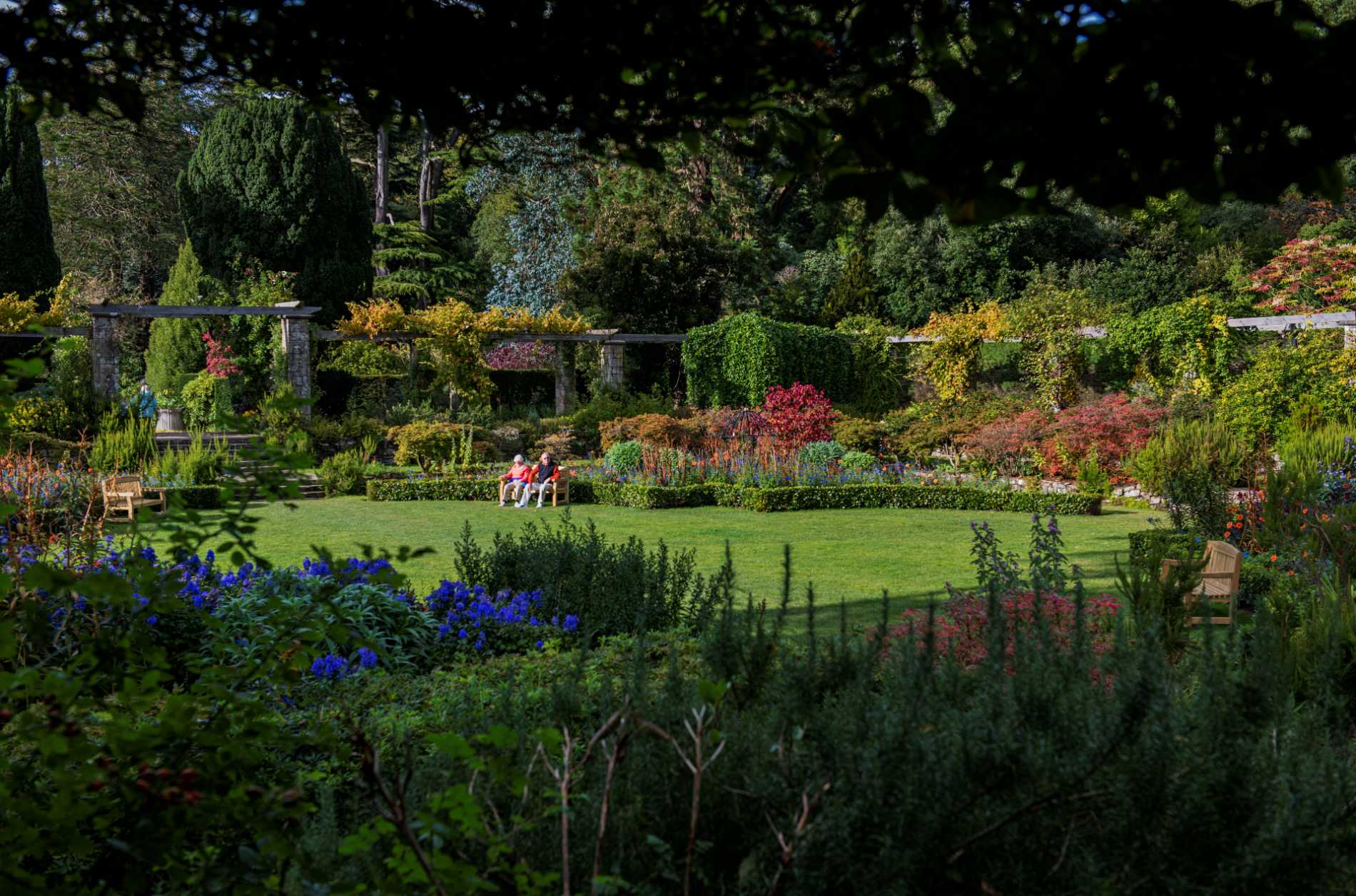 Sunlit gardens at Mount Stewart with colourful flowerbeds, manicured lawns, and visitors relaxing on benches.