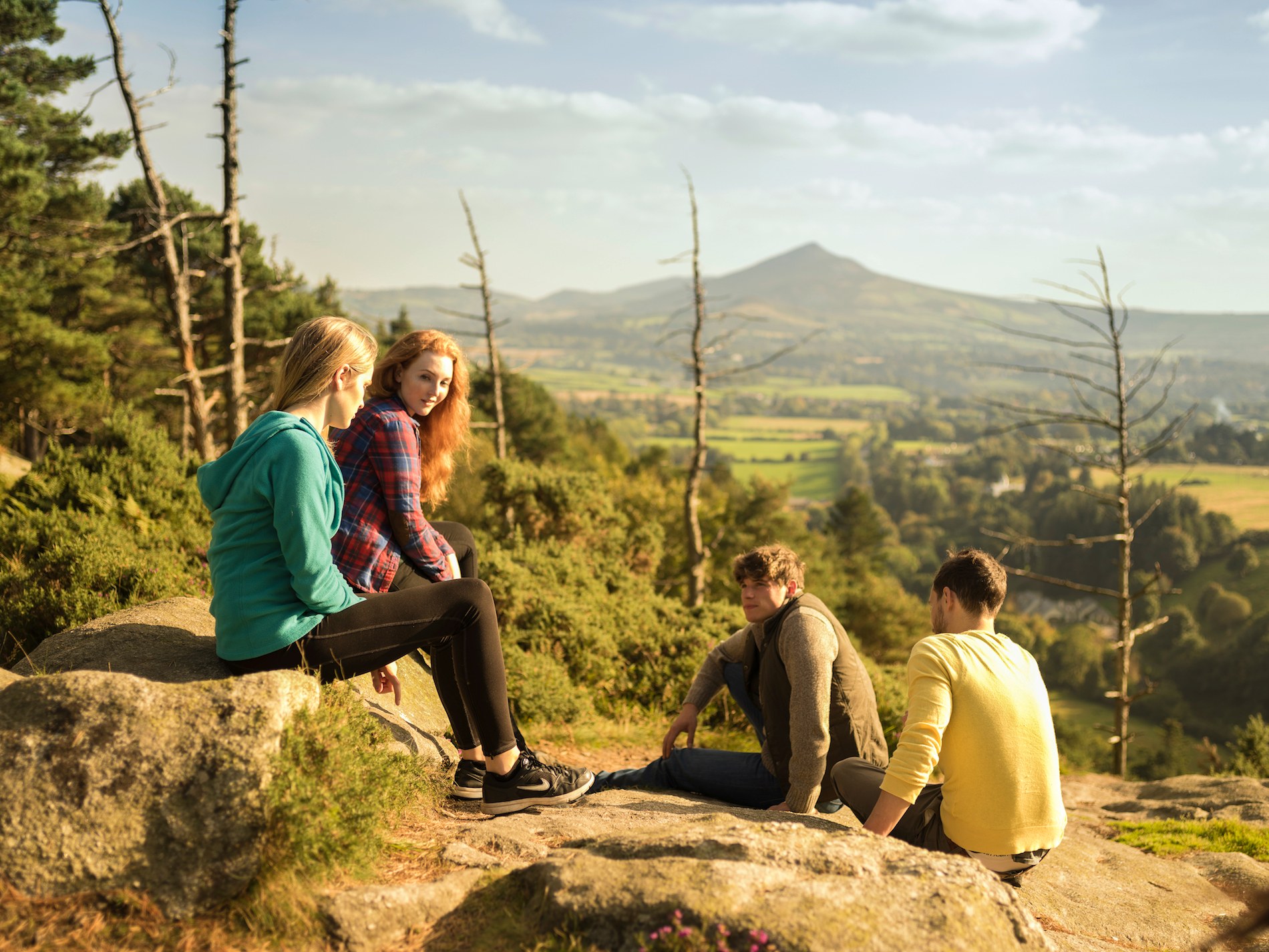 A group of young people sit chatting outdoors with a green valley backdrop.