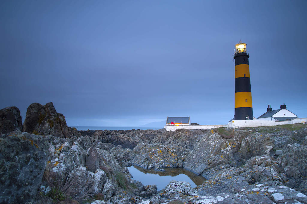 St John’s Point lighthouse with yellow and black stripes, set on rocky shoreline at dusk.