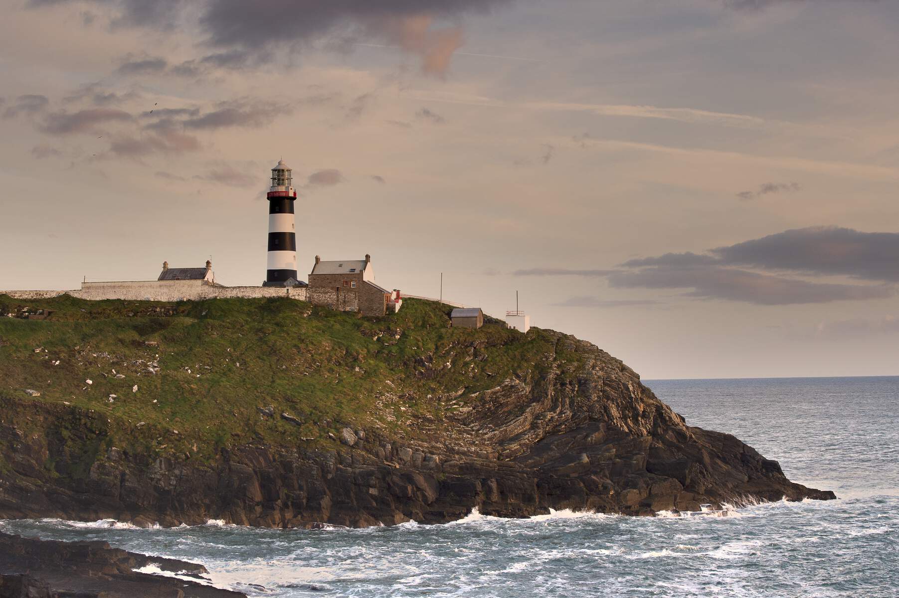 Black-and-white striped lighthouse on rocky headland at sunset, with cottages and sea below.