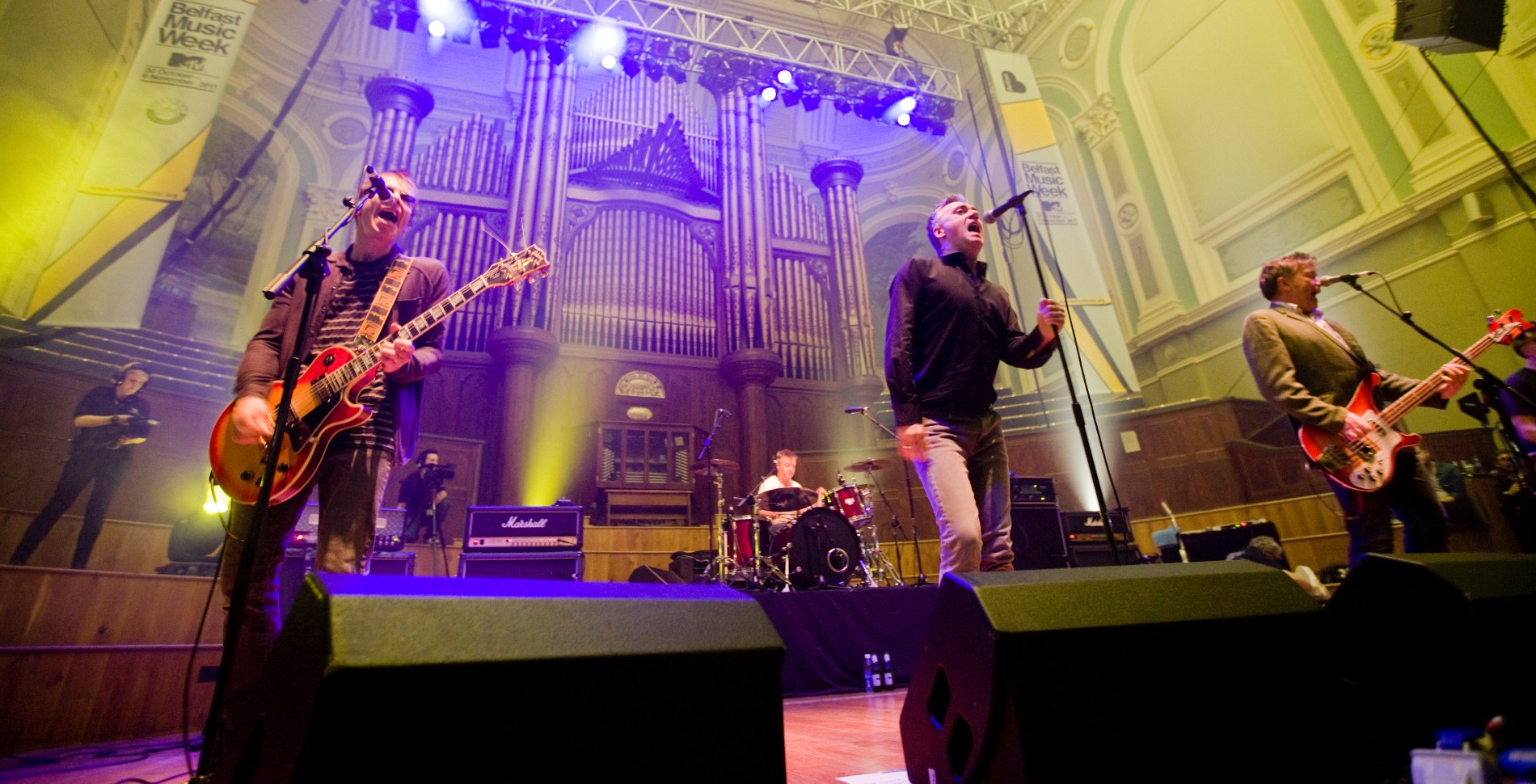 Energetic band plays live at a packed venue during Belfast Music Week, with dramatic lighting and a grand organ backdrop.