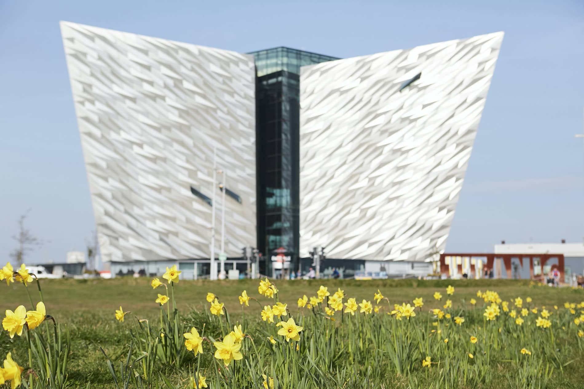 Titanic Belfast’s iconic ship-shaped building framed by daffodils under a bright spring sky.