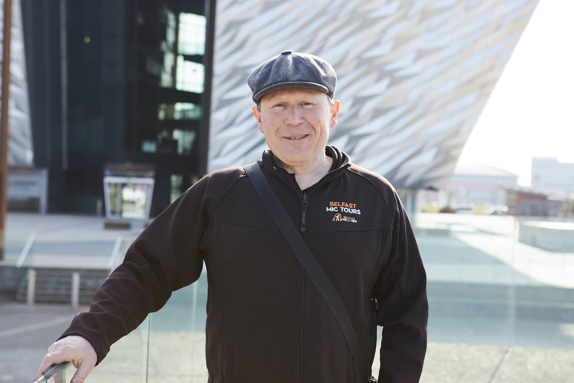 Tour guide Donal Kelly stands smiling in front of Titanic Belfast, wearing a flat cap and branded jacket.