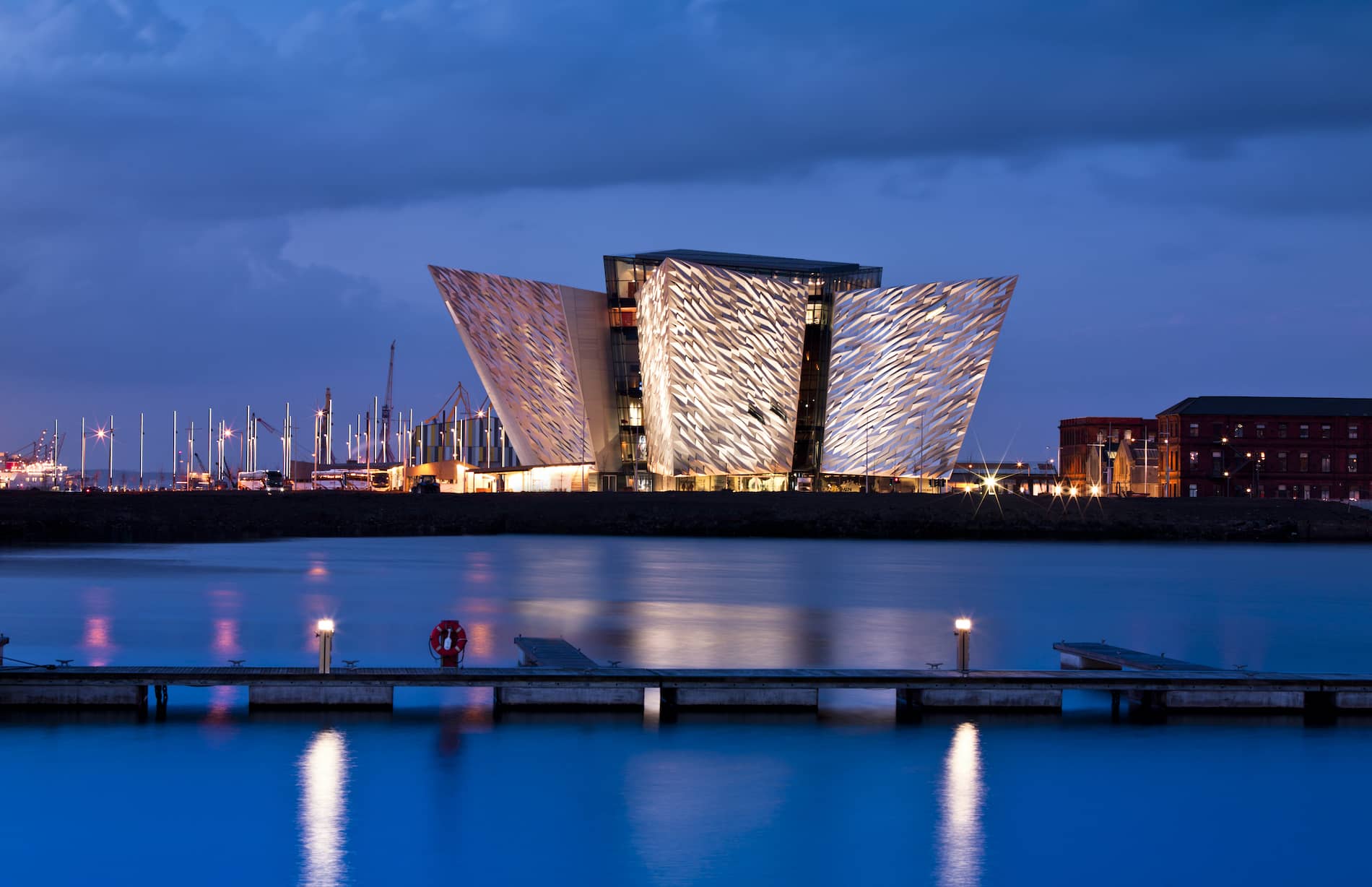 Titanic Belfast lit up at dusk, its angular facade reflected in the water, with a dark blue sky behind.