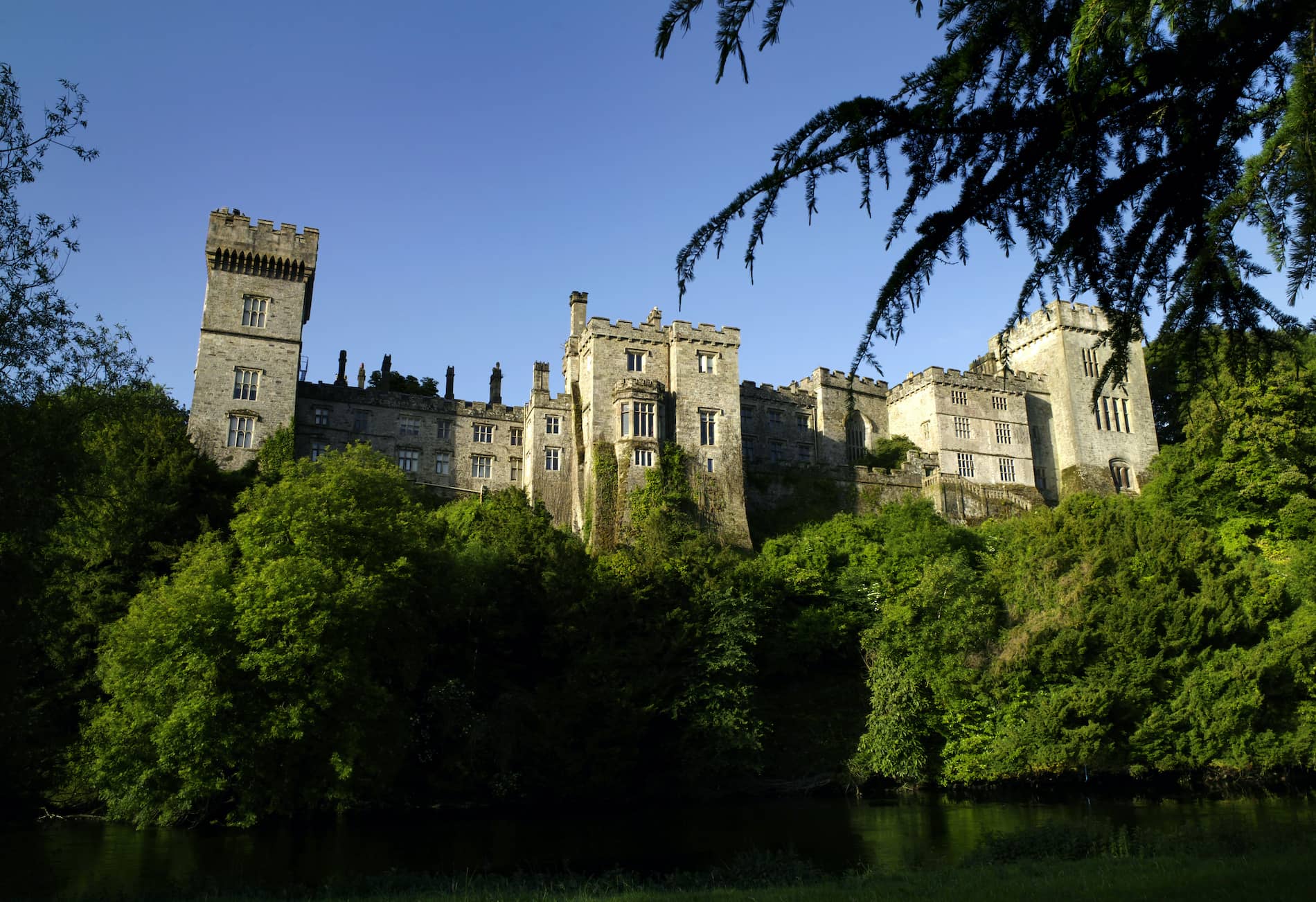 Lismore Castle with battlements rising above treetops and riverbank under a clear blue sky in Waterford.