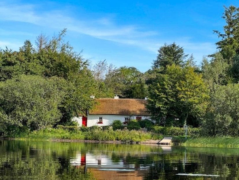 Whitewashed and thatched Keenaghan Cottage reflected in a calm lakeside setting surrounded by forest in Fermanagh.