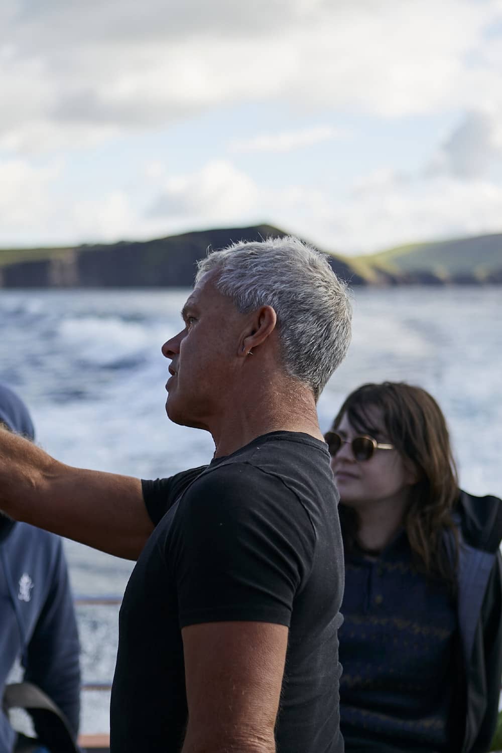 Male marine wildlife guide with short grey hair points toward the horizon while speaking to tourists on a boat off Dingle.