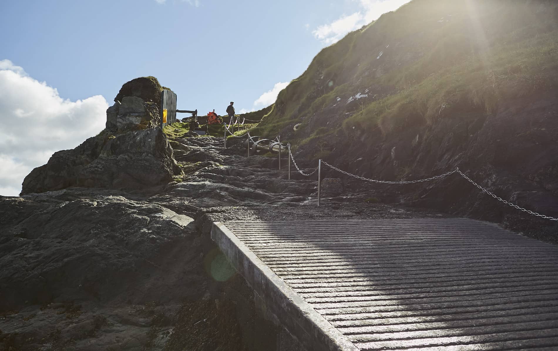 Steep landing and path leading up from the shore of Blasket Island, with sun flare above cliffs and safety chains in view.