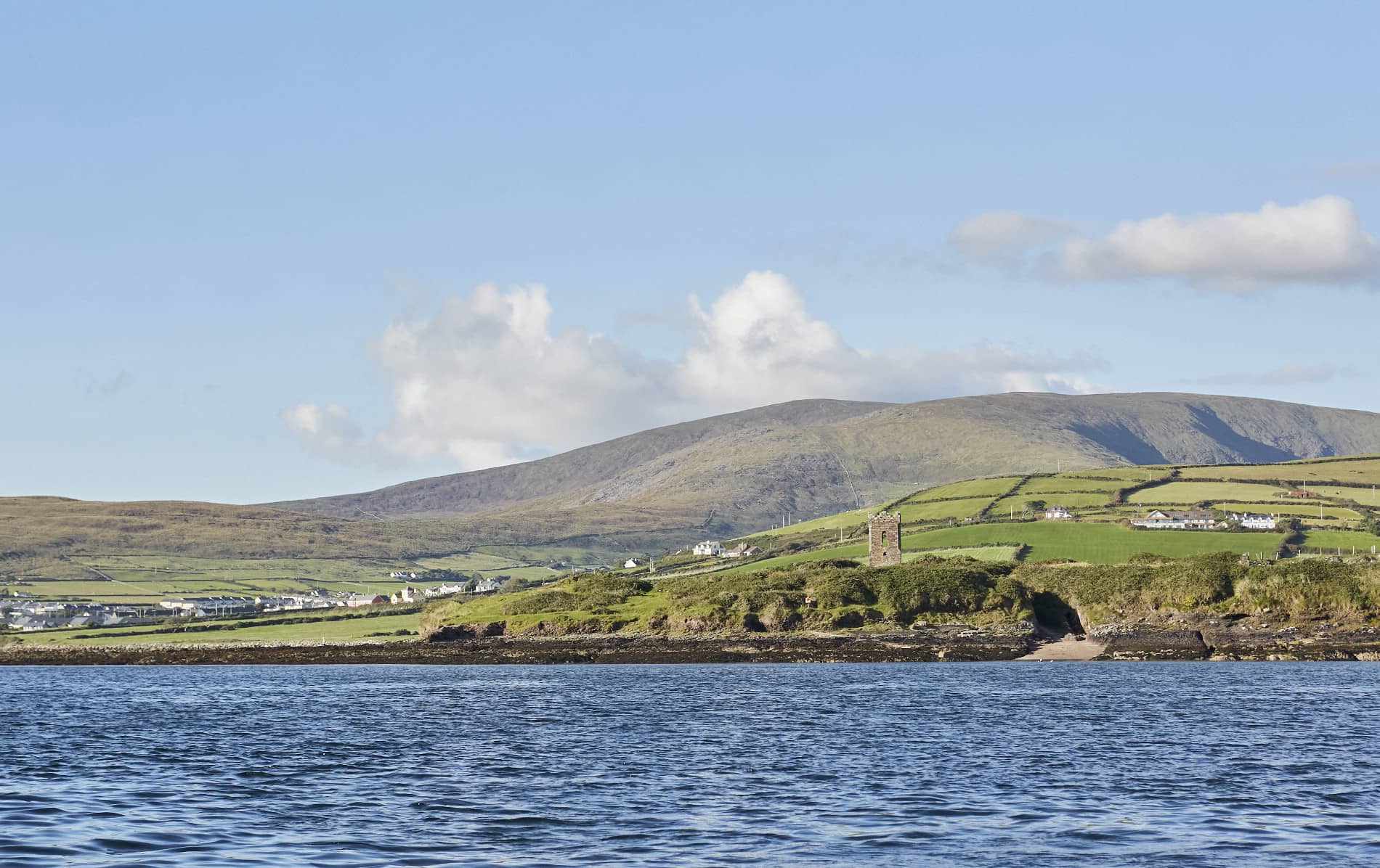 View of Ballydavid village with scattered houses and a medieval tower against the Slieve Mish mountains under a clear sky.