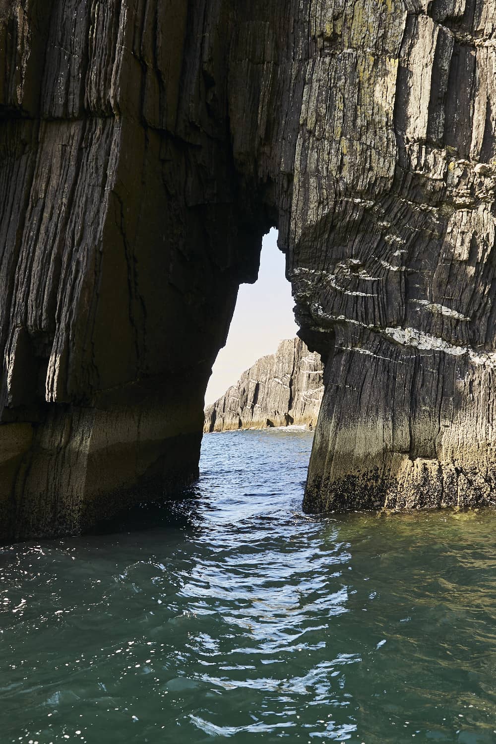Narrow sea arch cut through layered black cliffs, revealing clear blue water and more jagged rock formations beyond.