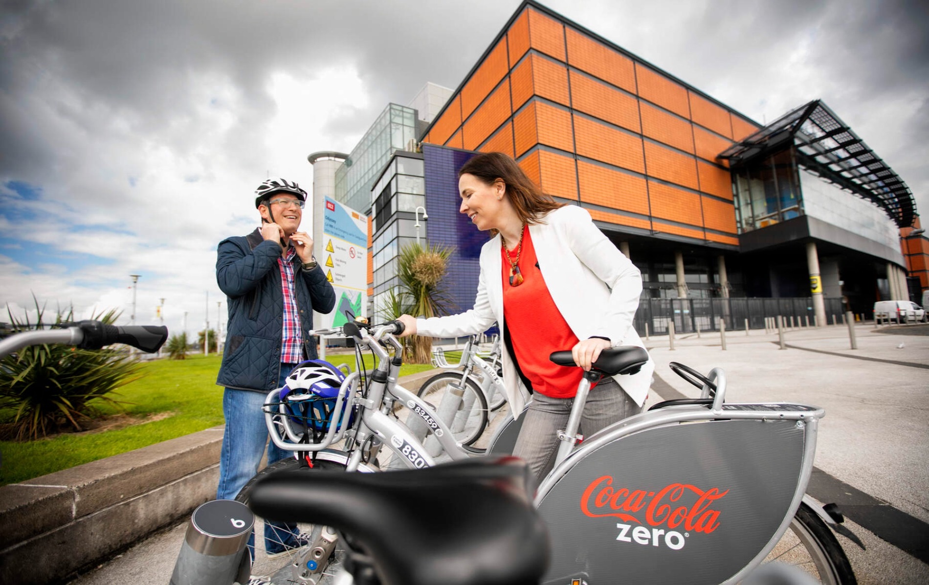 Two people prepare to cycle from a Belfast bike-share station outside the Odyssey Complex, promoting green urban travel.