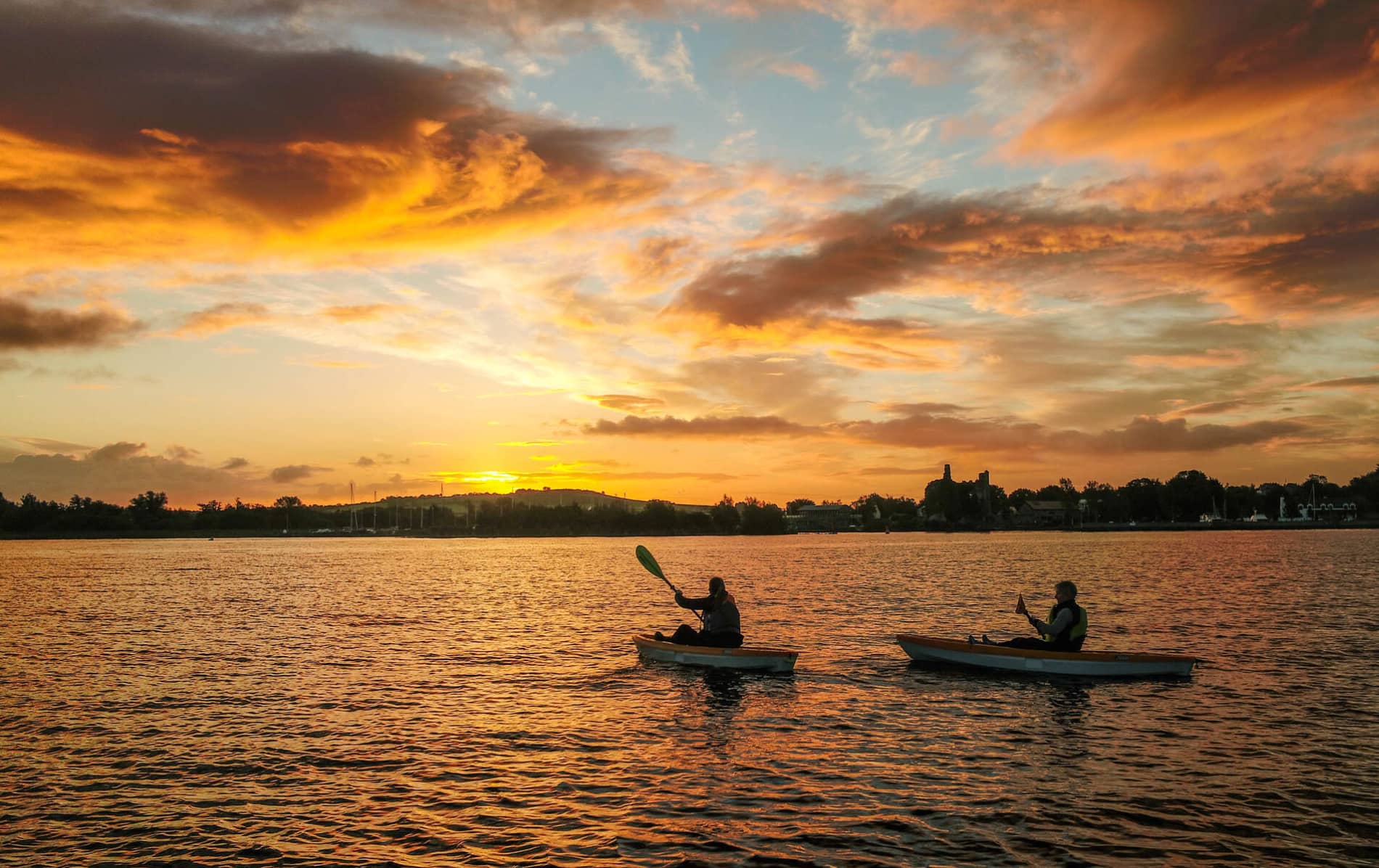 Two solo kayakers paddle across Lough Derg at sunset, silhouetted against a glowing sky and distant village shoreline.