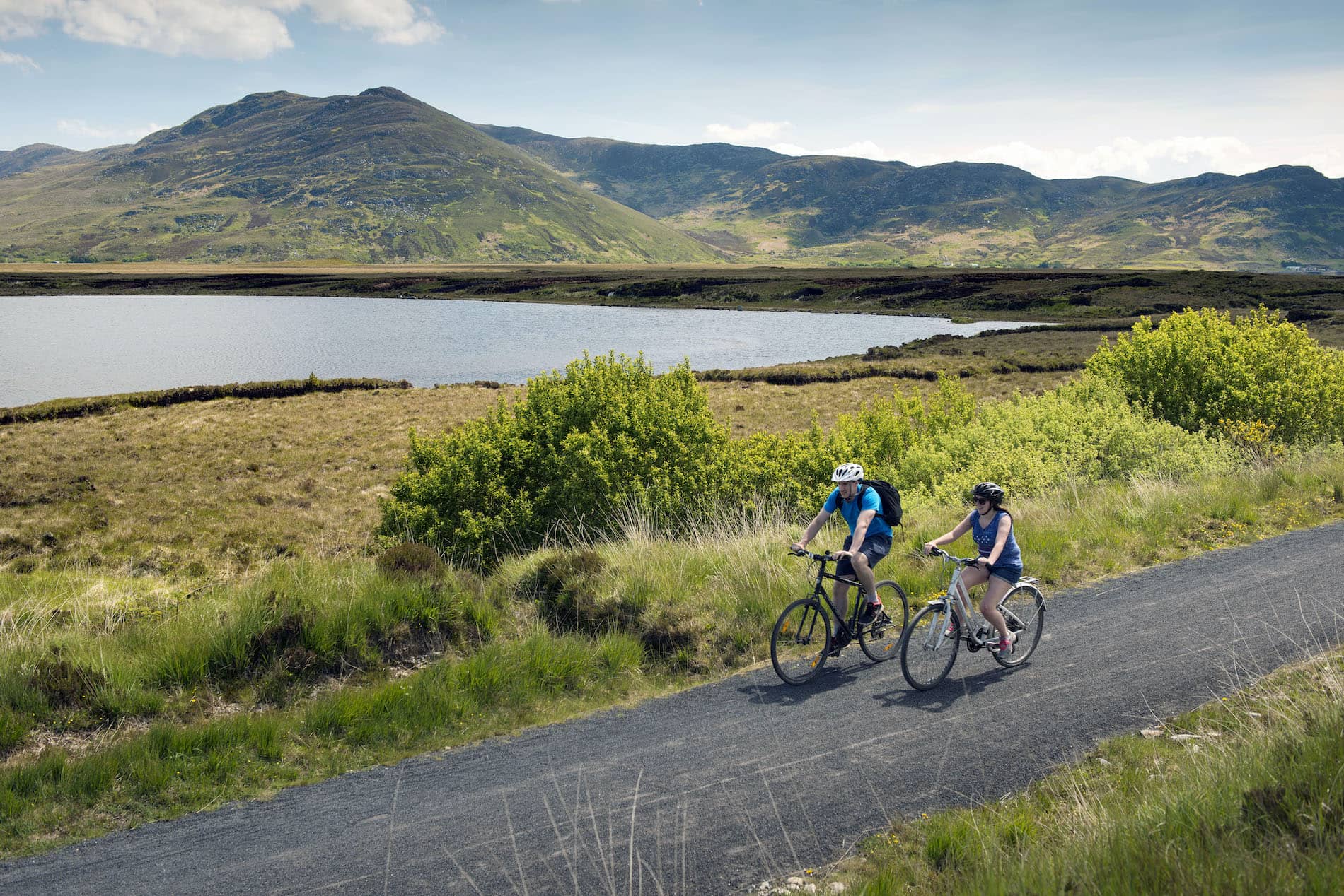 Cyclists ride the Great Western Greenway in County Mayo, past a lake and green hills under a clear sky.