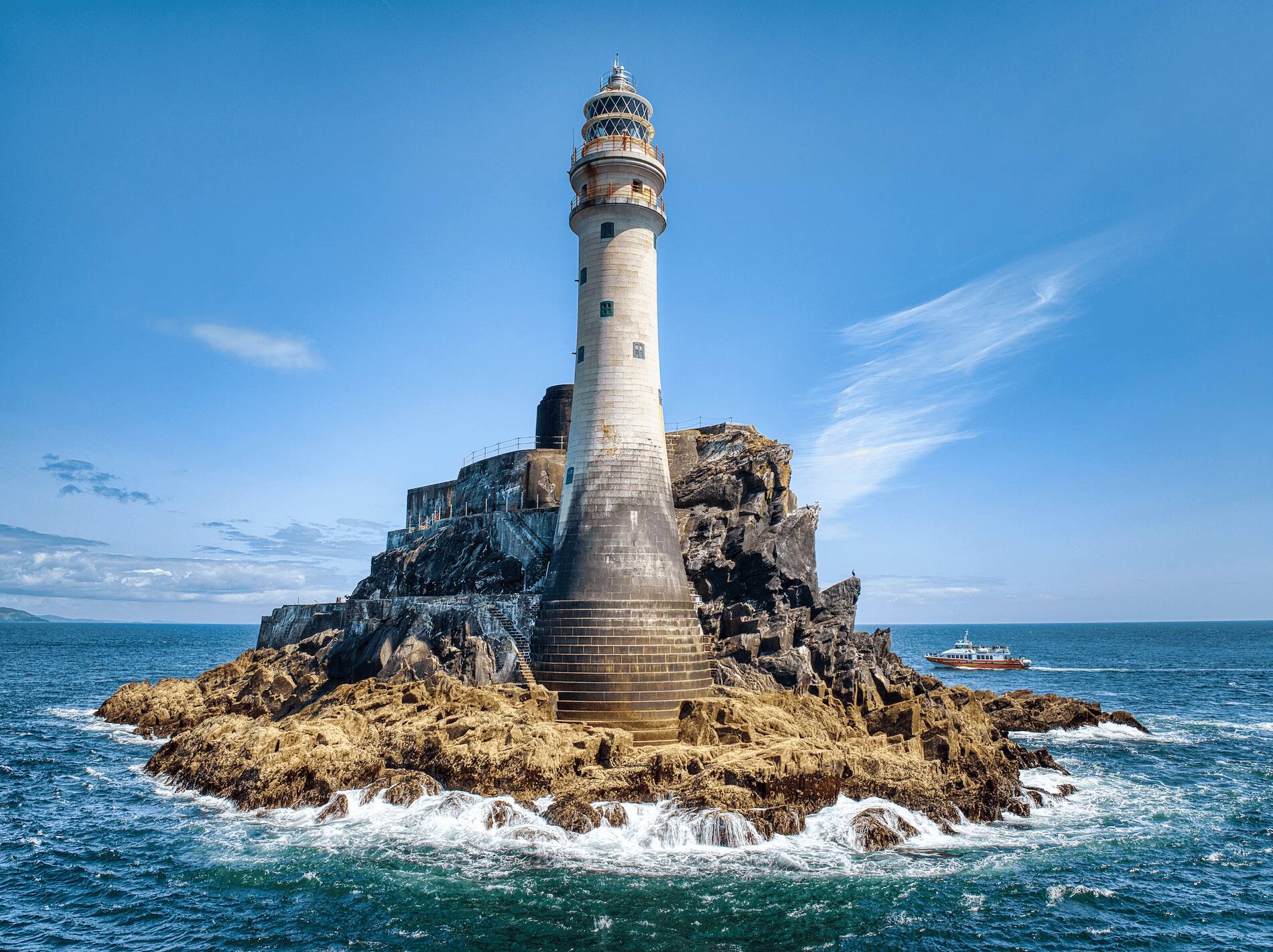 Fastnet Lighthouse rises from a rocky outcrop off the Cork coast, with waves crashing and a boat nearby.