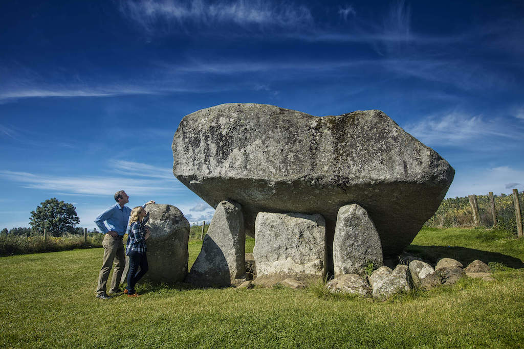 Two people standing beside Brownshill Dolmen in a grassy field under a deep blue sky in Carlow.