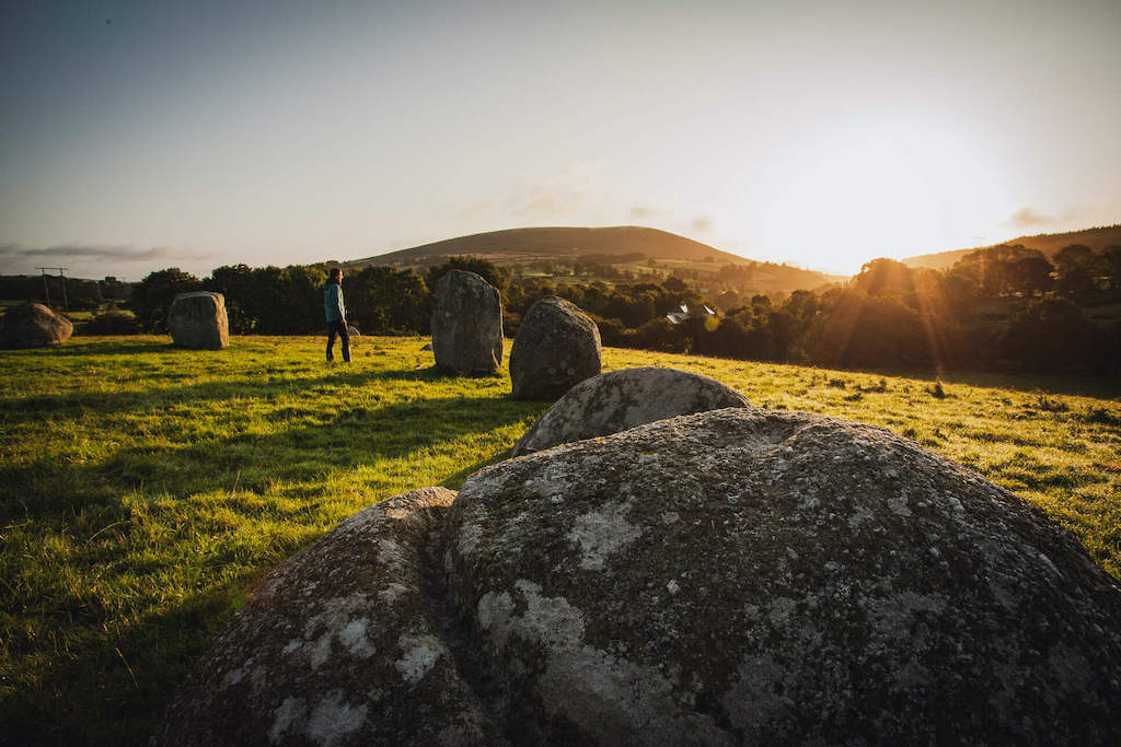 Person walking among granite boulders at Athgreany stone circle in Wicklow, with golden evening sun behind hills.