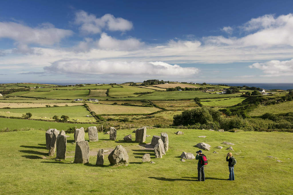 Two people looking at Drombeg stone circle on a grassy slope above patchwork farmland and sea in Cork.