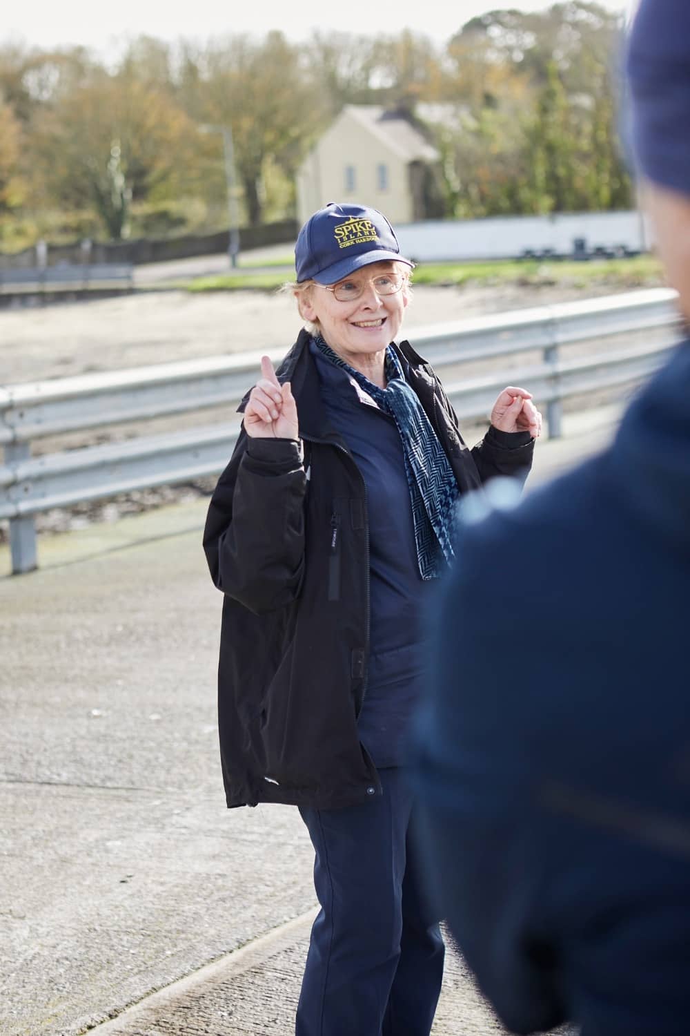 Smiling tour guide wearing Spike Island cap and jacket welcoming visitors.
