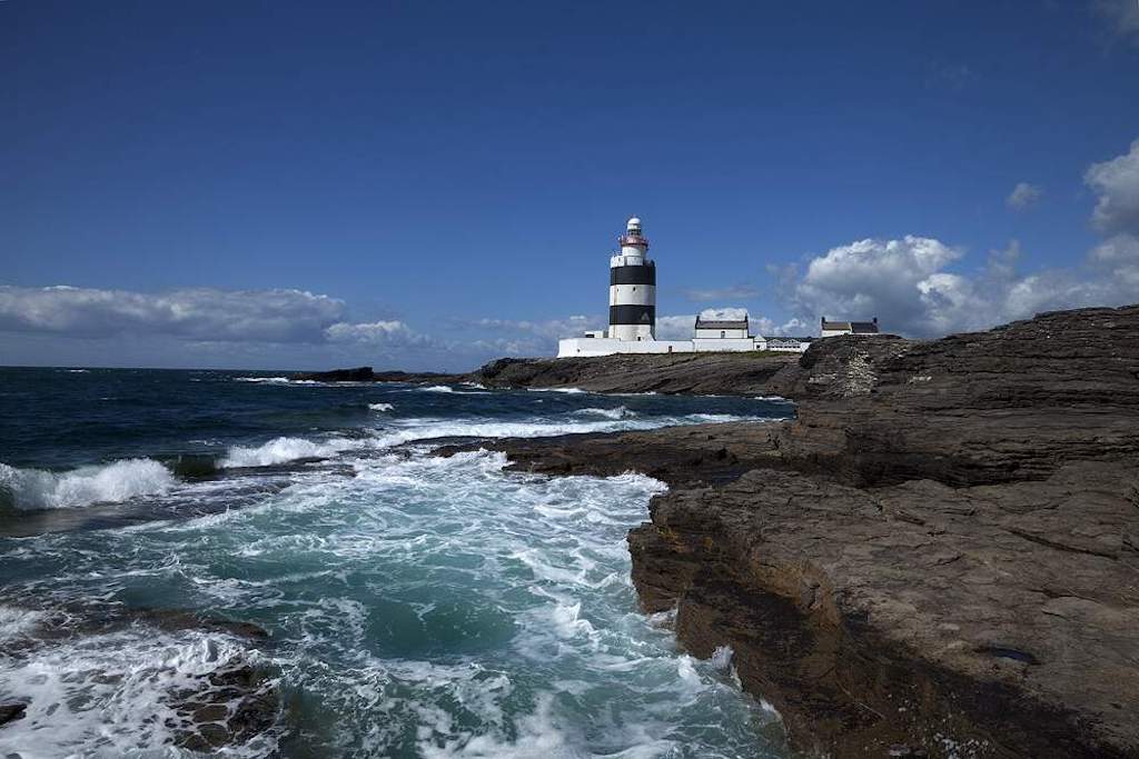 Hook Lighthouse on the Wexford coast with waves crashing against the rocky shore under a blue sky.