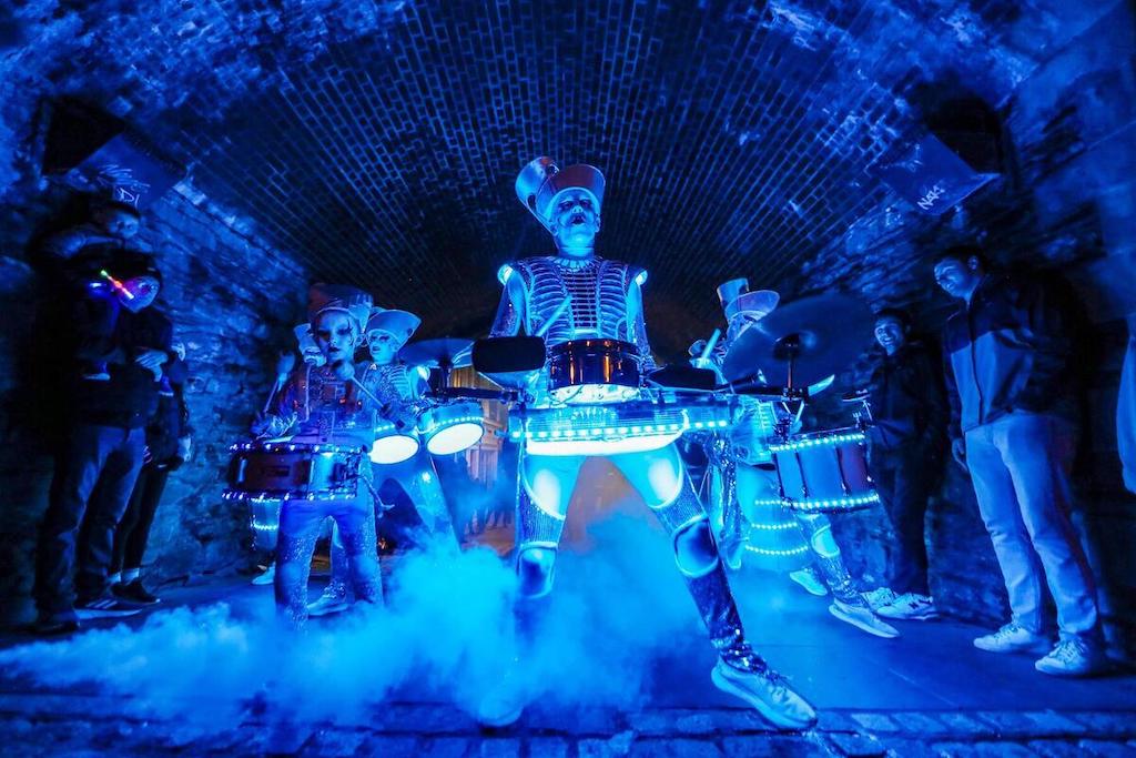 Costumed street performers playing illuminated drums during a Derry Halloween night event in a stone archway.