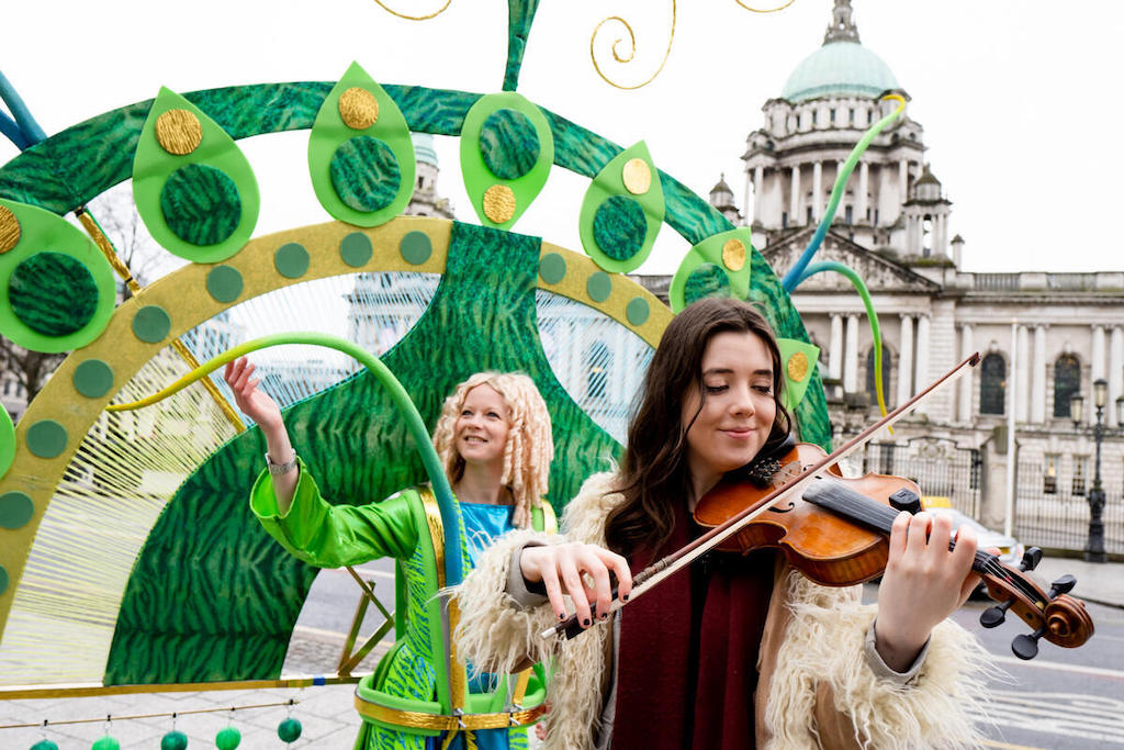 Musician playing a fiddle during a St Patrick’s Day celebration in Belfast, with colourful parade costumes and City Hall behind.