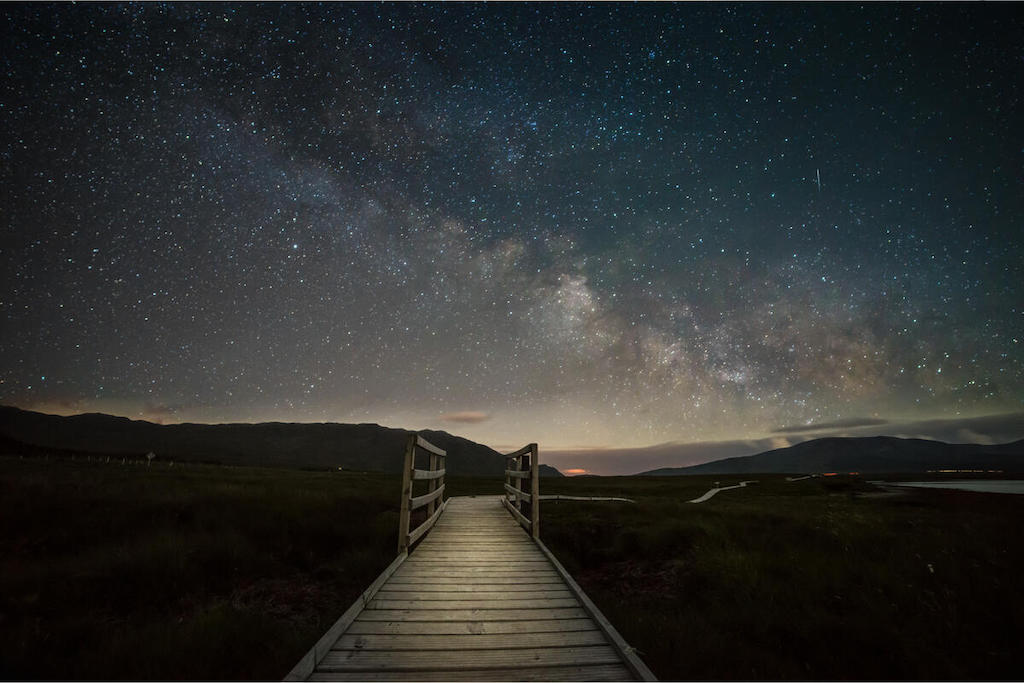 Wooden boardwalk leading into a County Mayo bog beneath a clear night sky filled with stars and the Milky Way.
