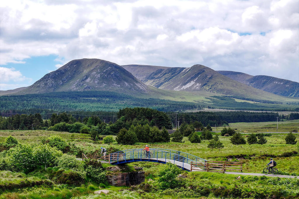 Cyclists crossing a bridge on the Great Western Greenway in County Mayo, with green fields and mountains beyond.