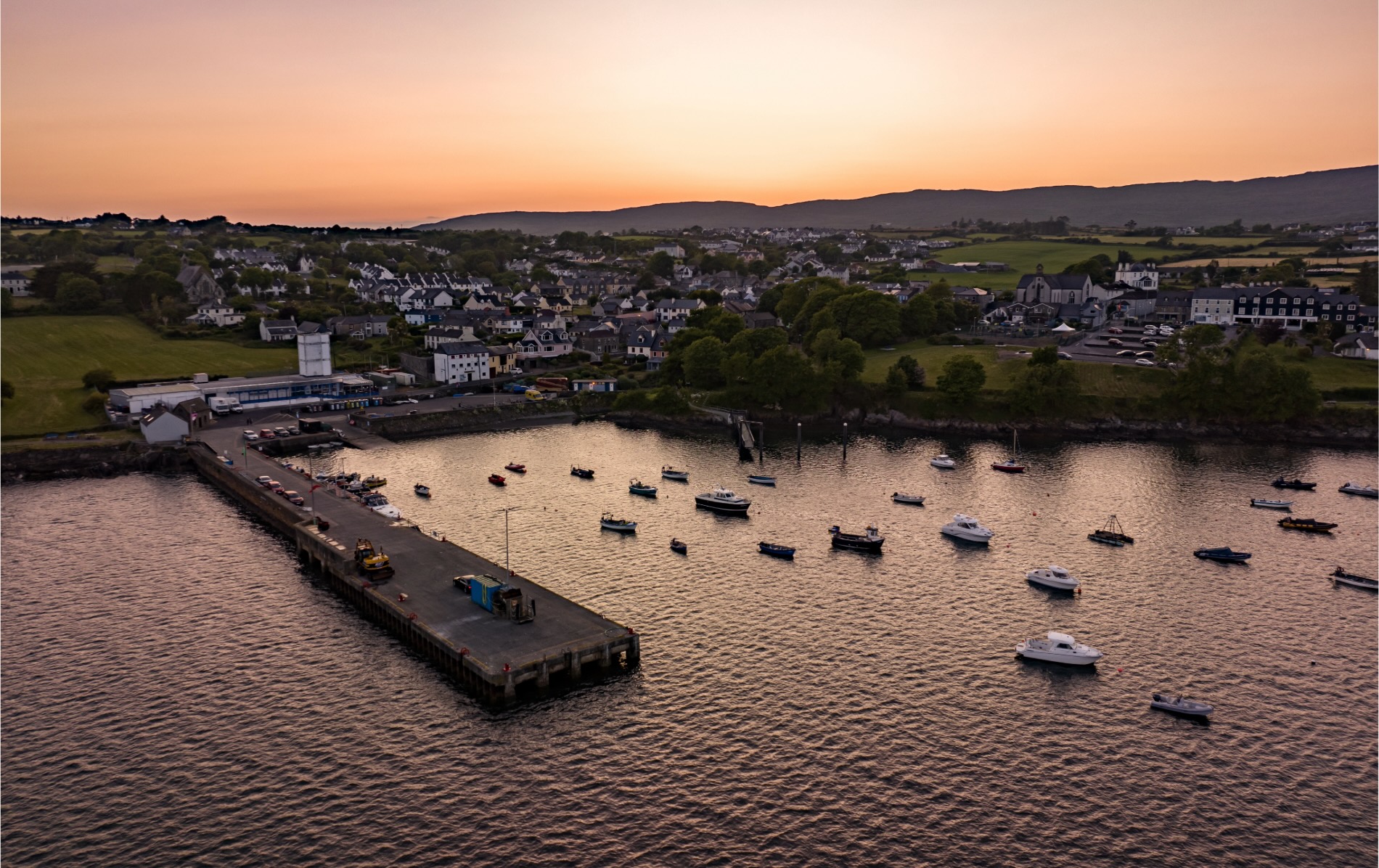 Sunset view over boats moored at Schull harbour in Cork with the town and hills in the background.