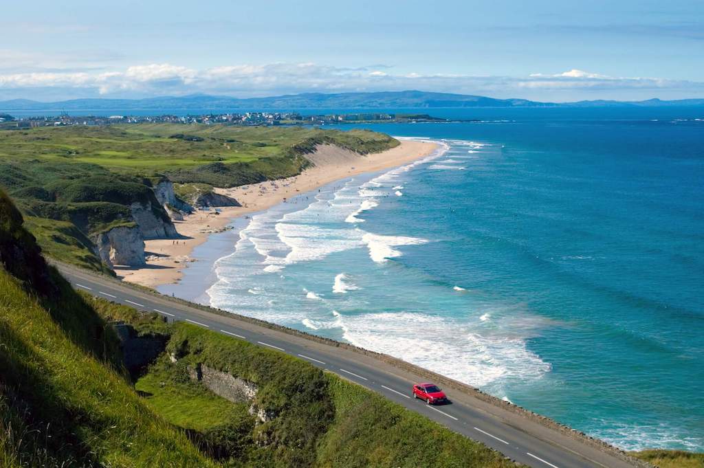 Red car driving the Causeway Coastal Route with sandy Whiterocks Beach and blue waves in Portrush, County Antrim.
