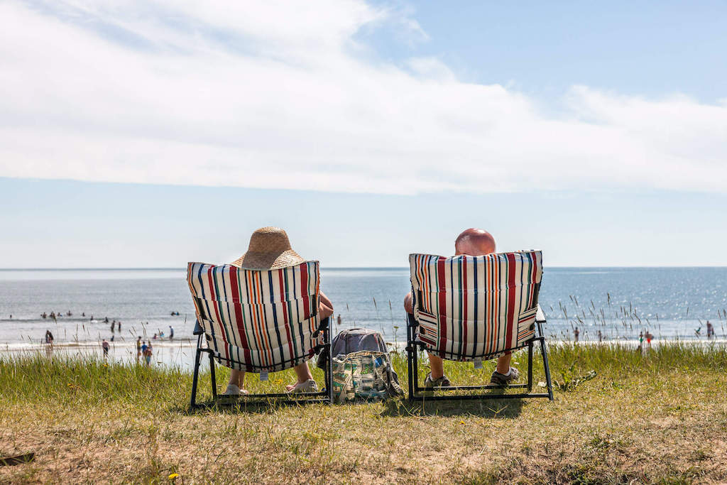 Two people relaxing on striped deck chairs overlooking a busy beach on a sunny summer day in Youghal, County Cork.