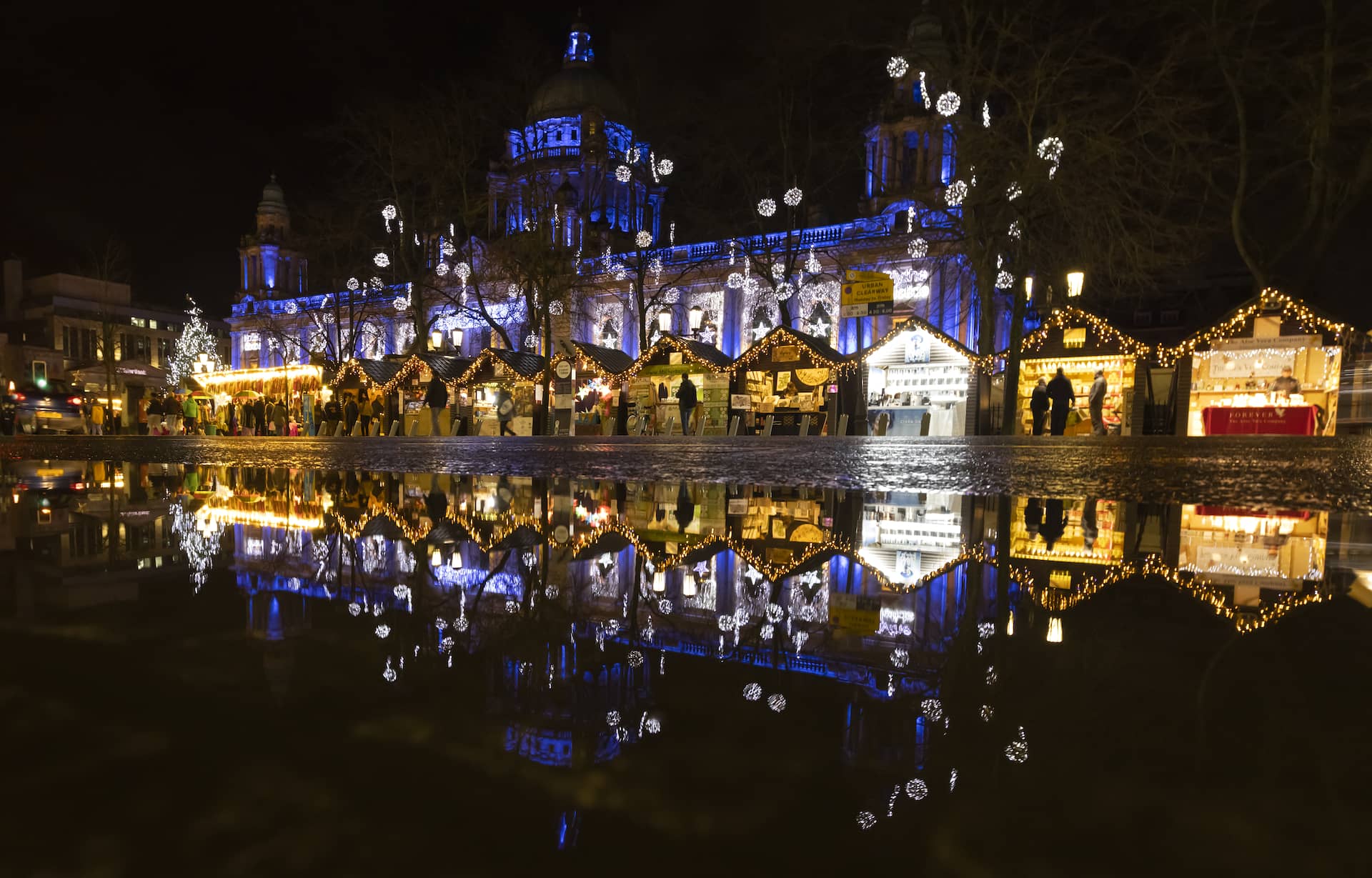 Christmas market huts glowing with fairy lights reflected in puddles before Belfast City Hall at night.