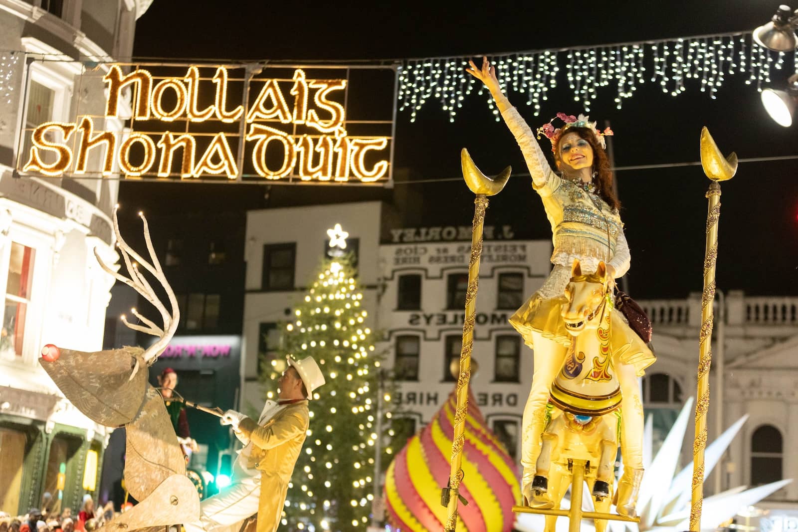 Corkmas festive parade in Cork city with performers on golden floats beneath a “Nollaig Shona Duit” sign and a lit Christmas tree in the background.