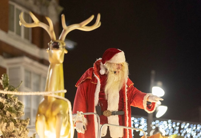 Santa in a red suit waving from a festive float beside a golden reindeer during a Christmas celebration.