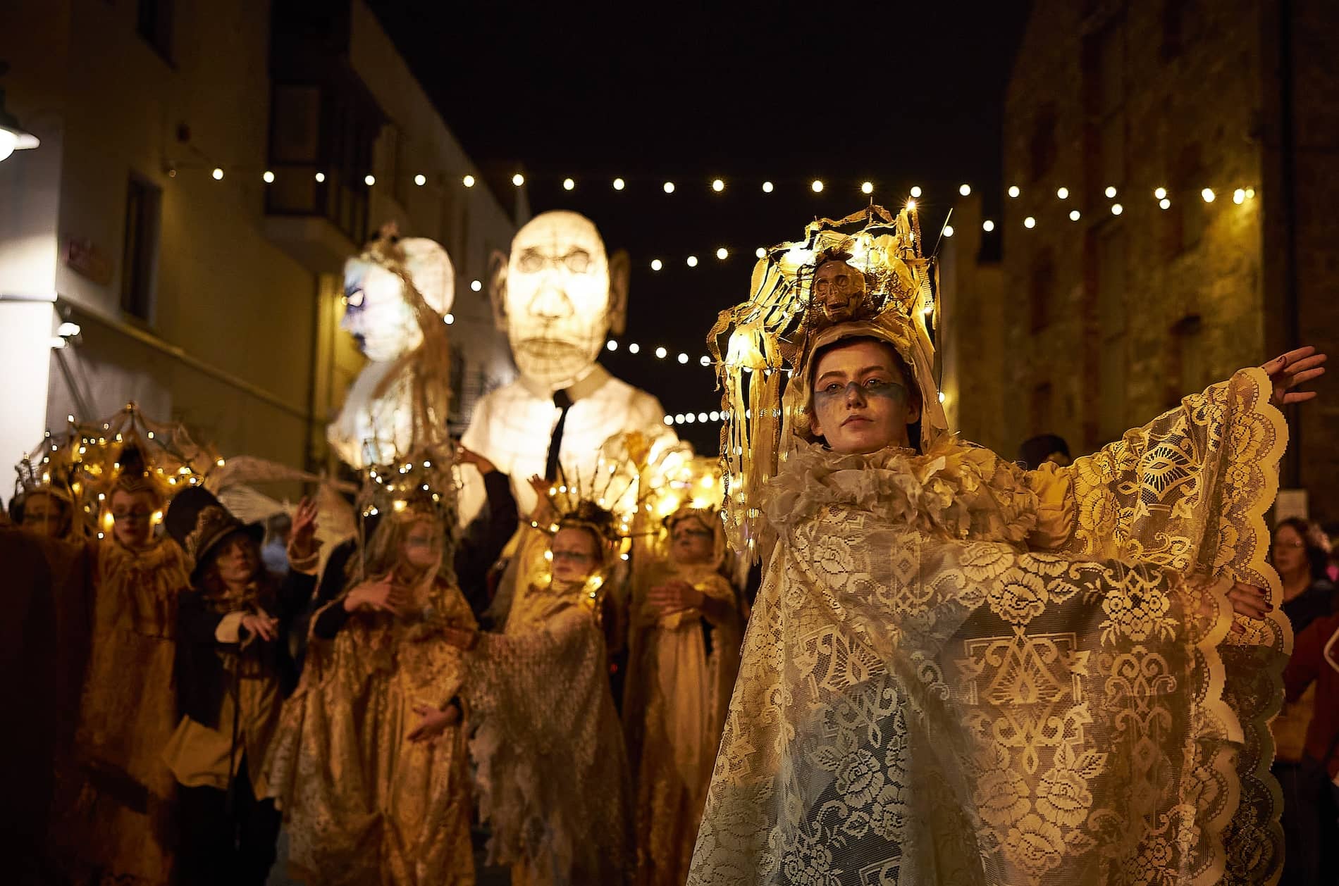 Performer in ornate lace costume with glowing headpiece leads night-time Halloween parade with giant lantern puppets in Limerick.