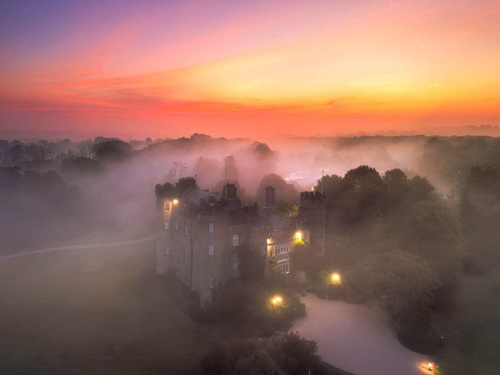 Malahide Castle surrounded by morning mist with glowing windows at sunrise in County Dublin.