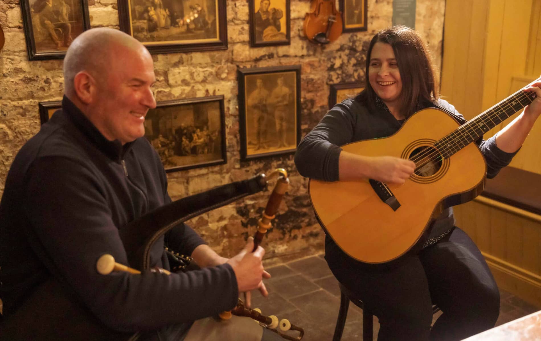 Two musicians play Irish traditional music on uilleann pipes and guitar in a cosy pub setting.