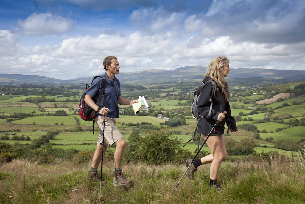 Two hikers with walking poles cross a grassy hill overlooking rolling countryside in the Wicklow Mountains.
