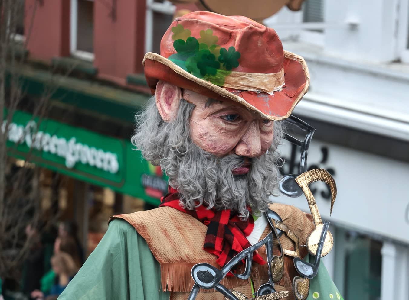 Costumed street performer with a large mask, shamrock hat and musical notes at a St Patrick’s Day festival in Enniskillen.