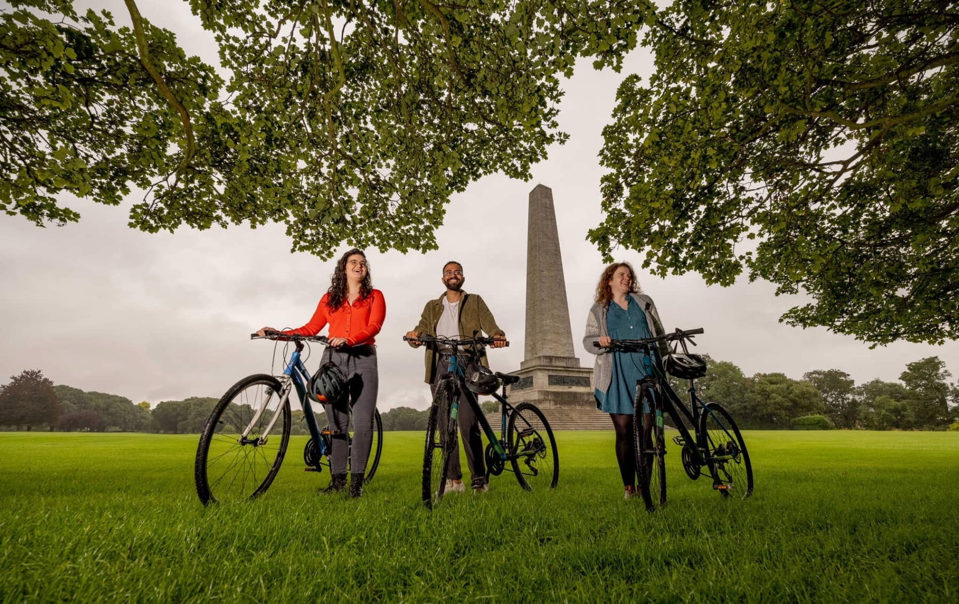 Three people with bicycles in Phoenix Park, Dublin, with the Wellington Monument in view.