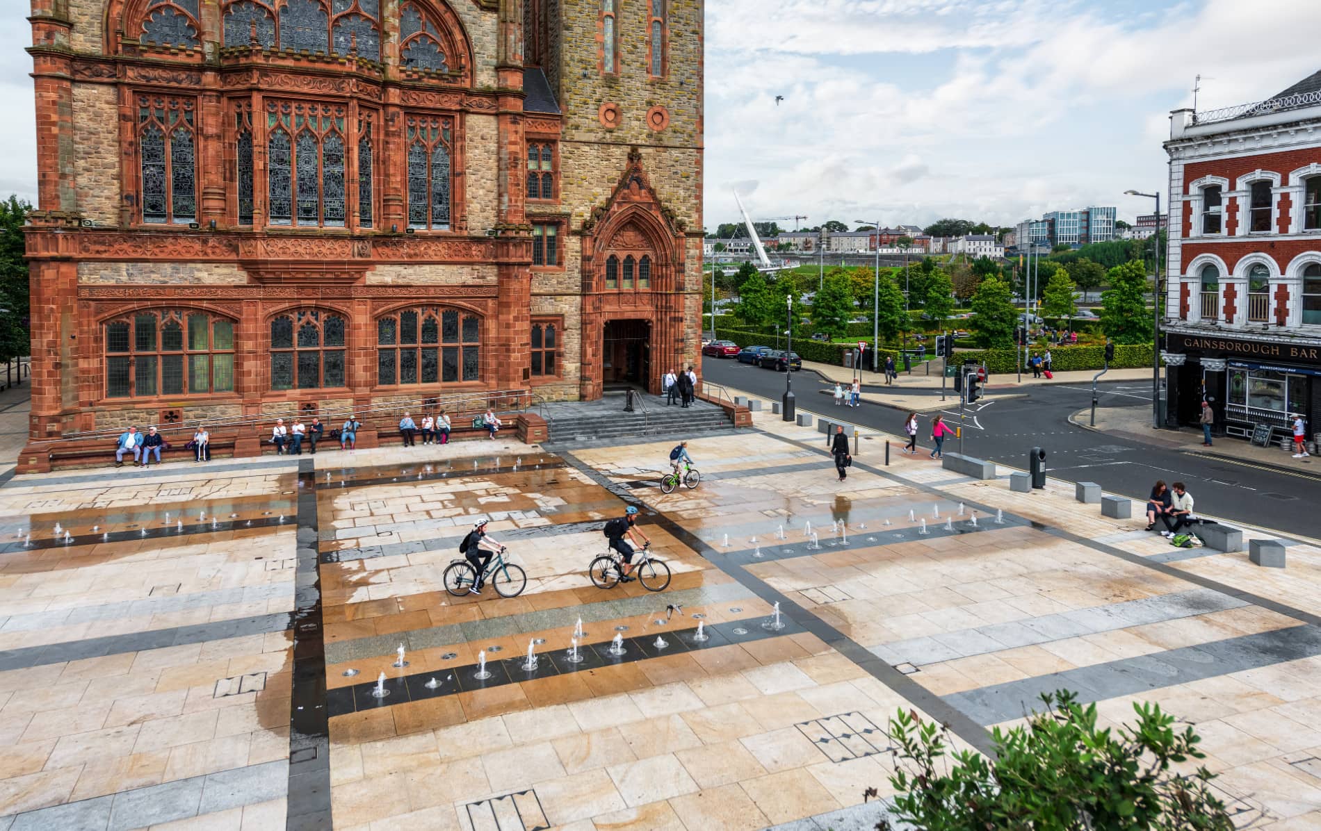 Cyclists riding across Guildhall Square in Derry~Londonderry with fountains and red-brick Guildhall.
