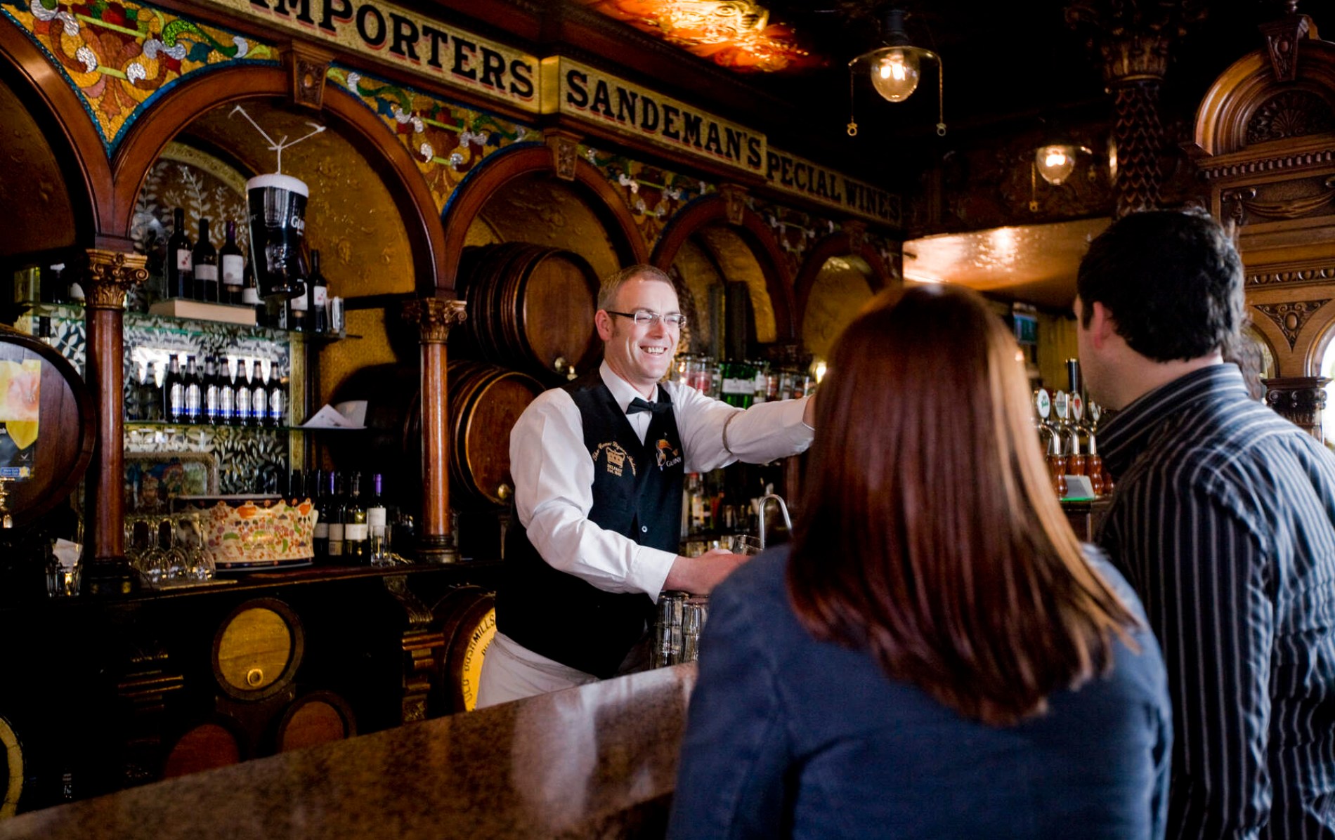 Smiling bartender serving drinks in a traditional Irish pub with ornate décor.