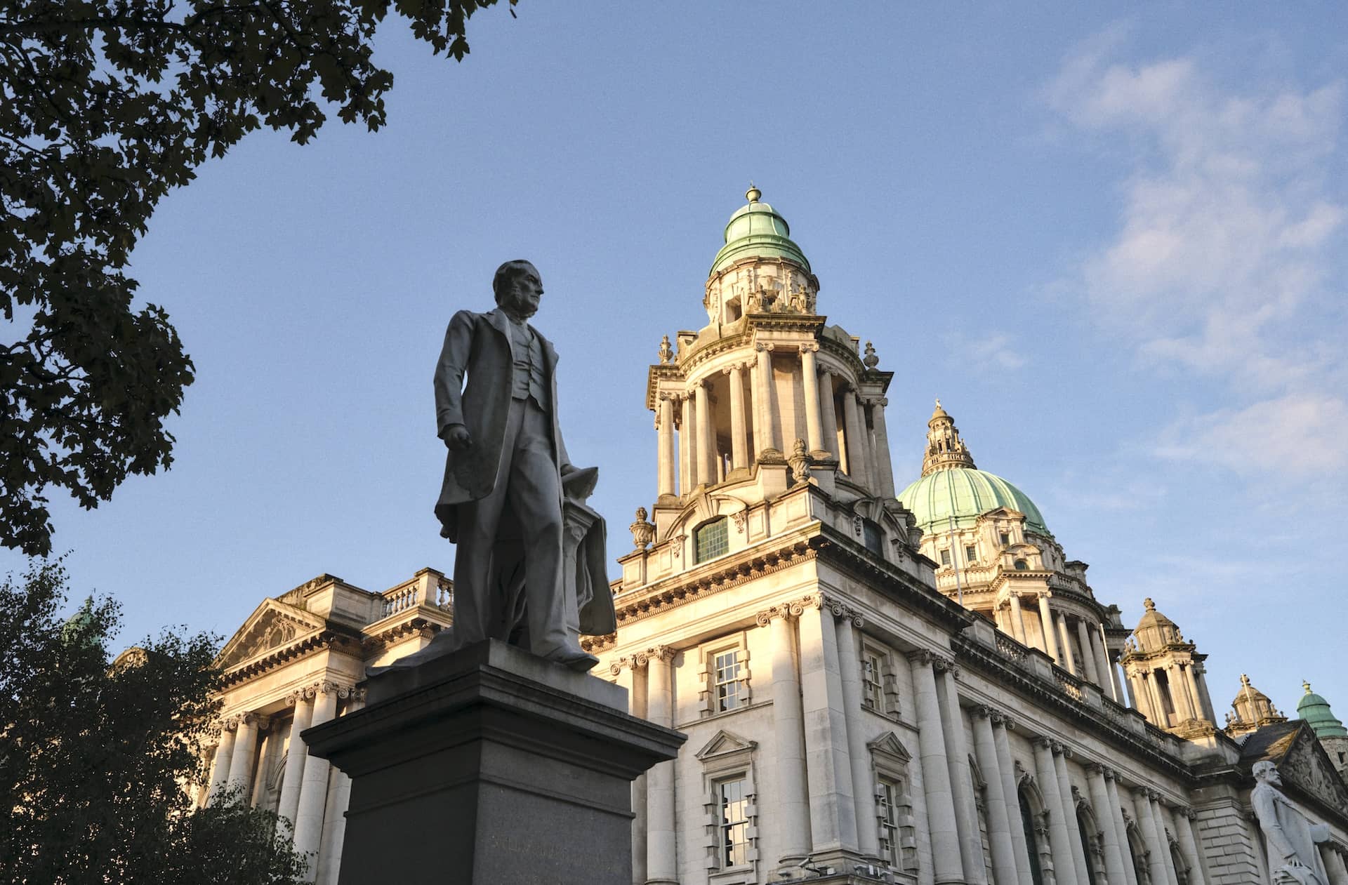 A statue stands before the grand domes and ornate façade of Belfast City Hall under a clear evening sky.
