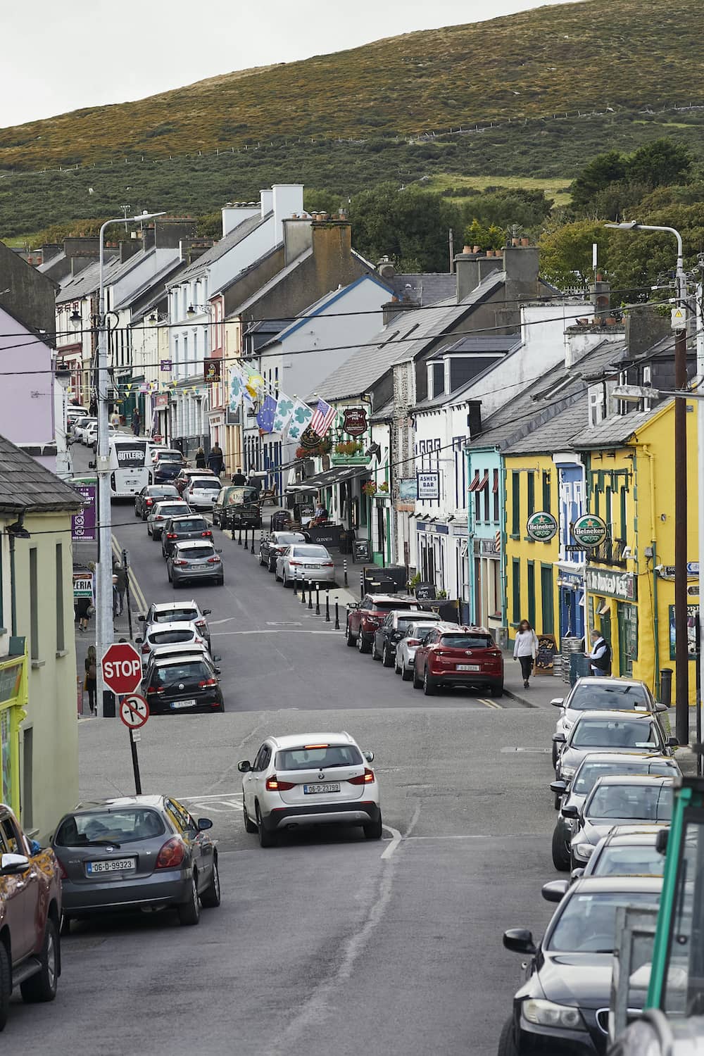 Busy main street in Dingle, lined with colourful pubs, shops, and parked cars against a hillside backdrop.